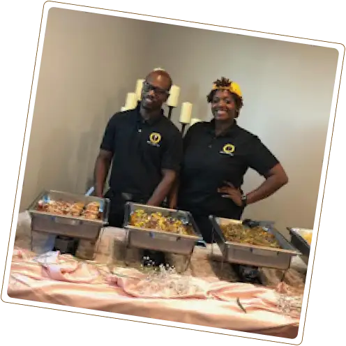 A man and a woman, both wearing black polo shirts with a logo, stand behind a catering table with three chafing dishes filled with food.