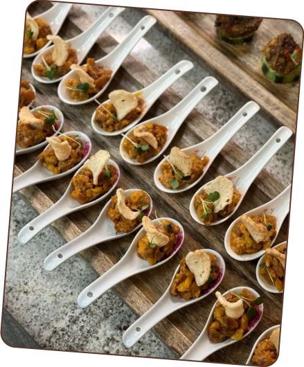 A diagonal, elevated view of multiple white ceramic spoons, each holding a small portion of an appetizer topped with a crisp and a garnish, arranged on a wooden serving tray.
