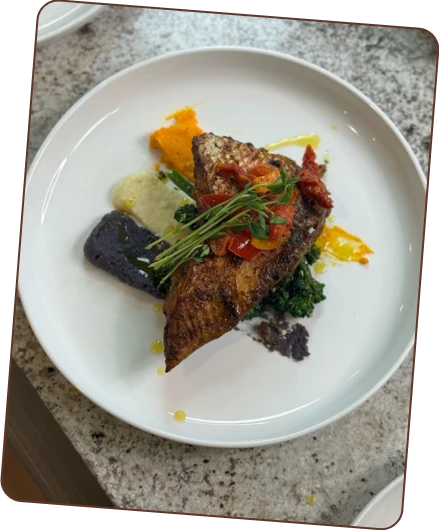 Plated dish with pan-seared fish, tomato-herb topping, colorful purees, dark greens, and sprouts.