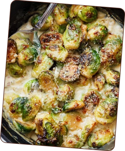 A close-up of roasted Brussels sprouts coated in a creamy sauce, sprinkled with cheese, and served in a dish with a spoon.