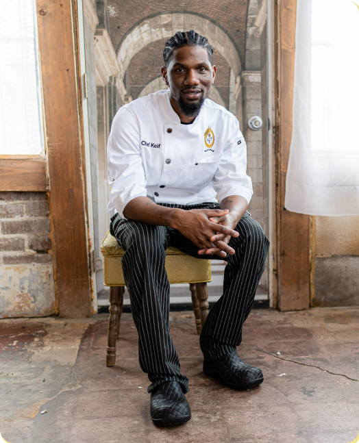 A male chef Keif with dreadlocks, wearing a white chef's coat and pinstriped pants, sits on a stool and smiles at the camera.